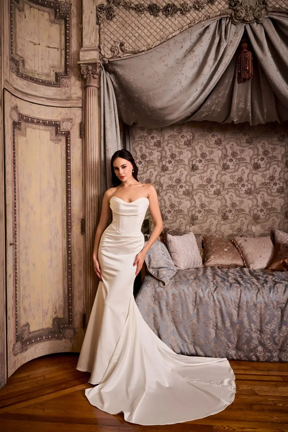 A woman in a fitted white gown stands beside a luxurious bed with ornate decor, under soft lighting.