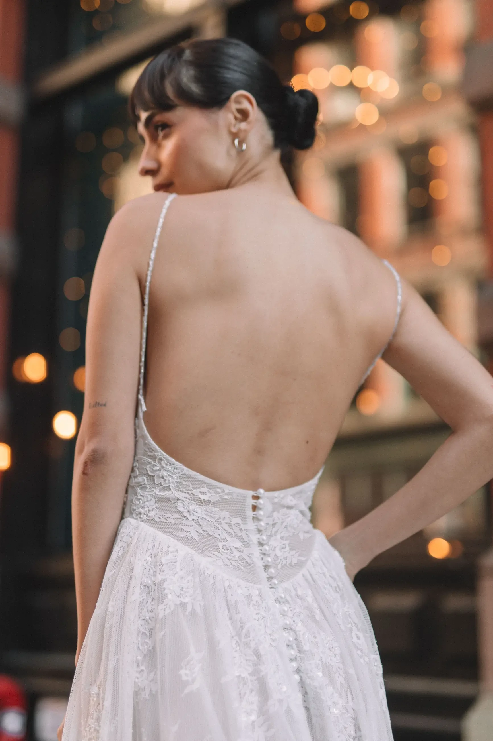 A woman in a backless wedding dress stands with her back turned towards the camera, showcasing the dress's intricate detailing and buttons, set against an urban backdrop with soft lighting.