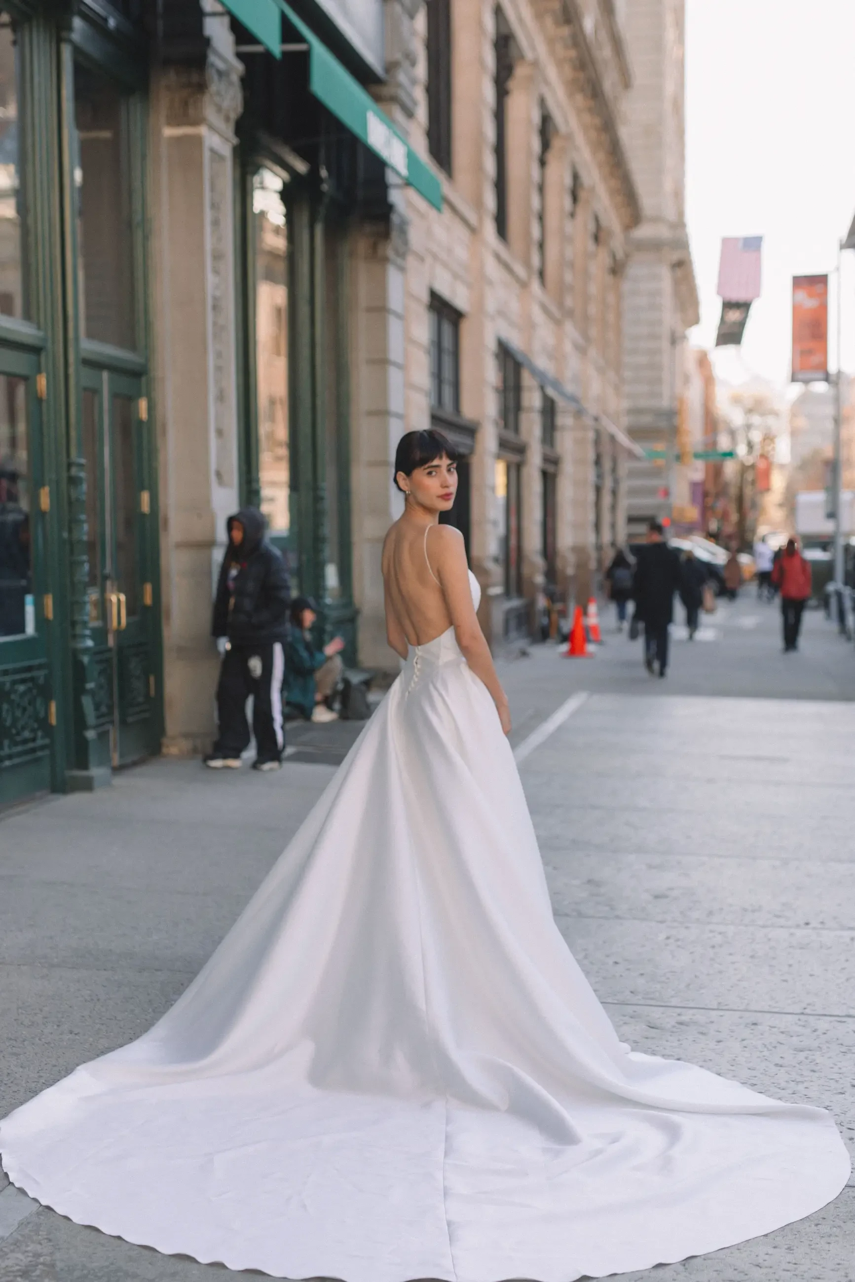 A woman wearing a white gown stands on a city street, looking over her shoulder. The gown has a long train and an open back. City buildings are visible in the background, along with pedestrians.