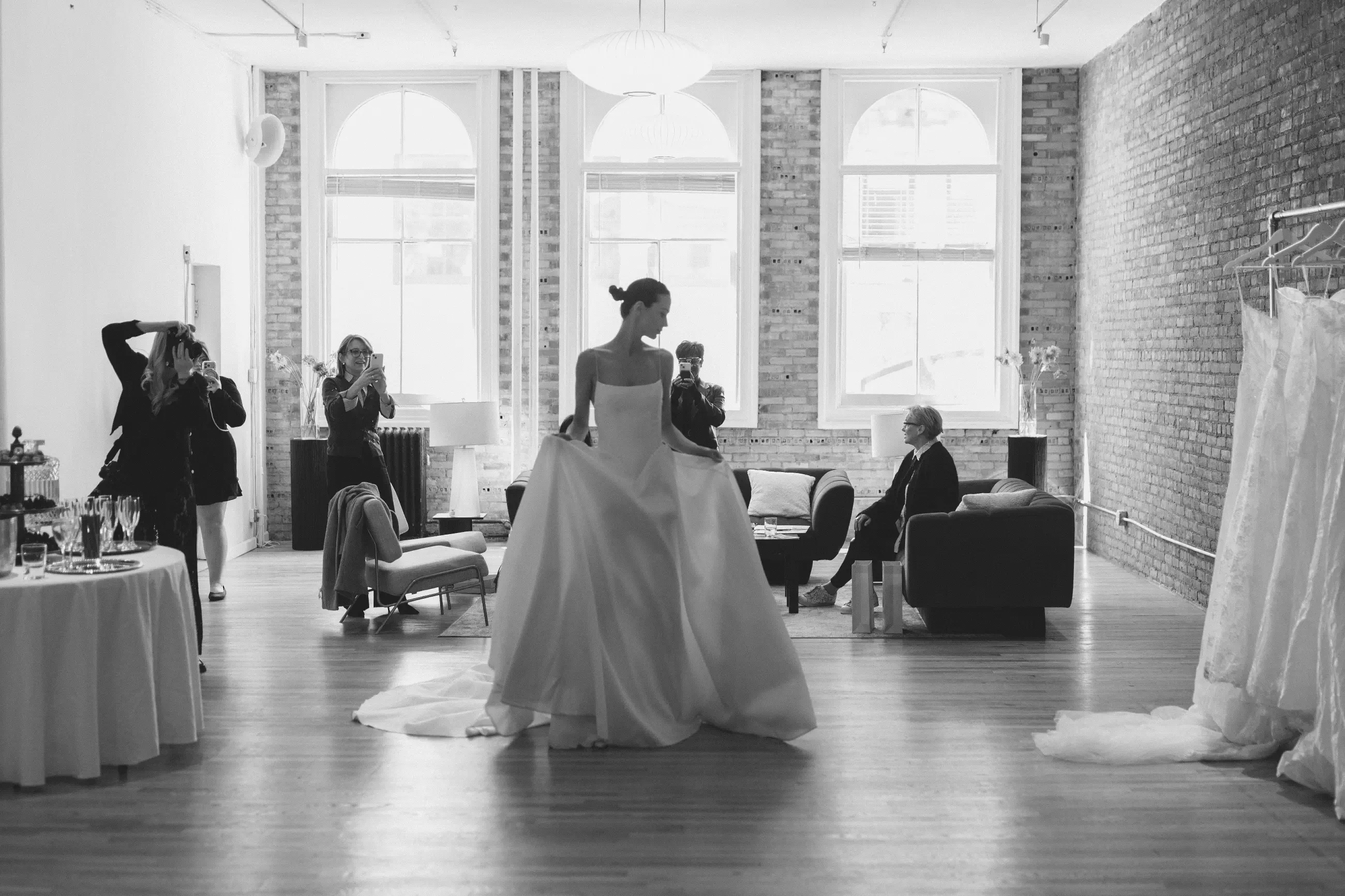 A bride in a wedding gown stands in a spacious room with large windows, while several people are engaged in various activities around her.