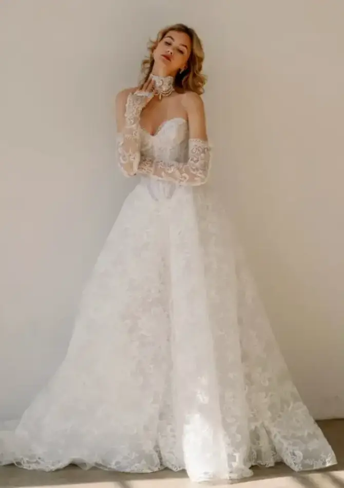 A woman in a strapless wedding dress with lace details and long lace gloves, posing elegantly against a plain background.