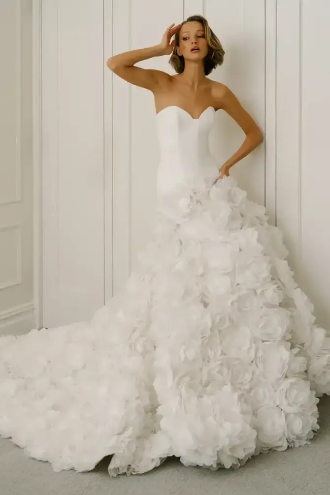 A model wearing a strapless wedding dress adorned with large white floral appliqu&eacute;s, posing gracefully against a neutral backdrop.