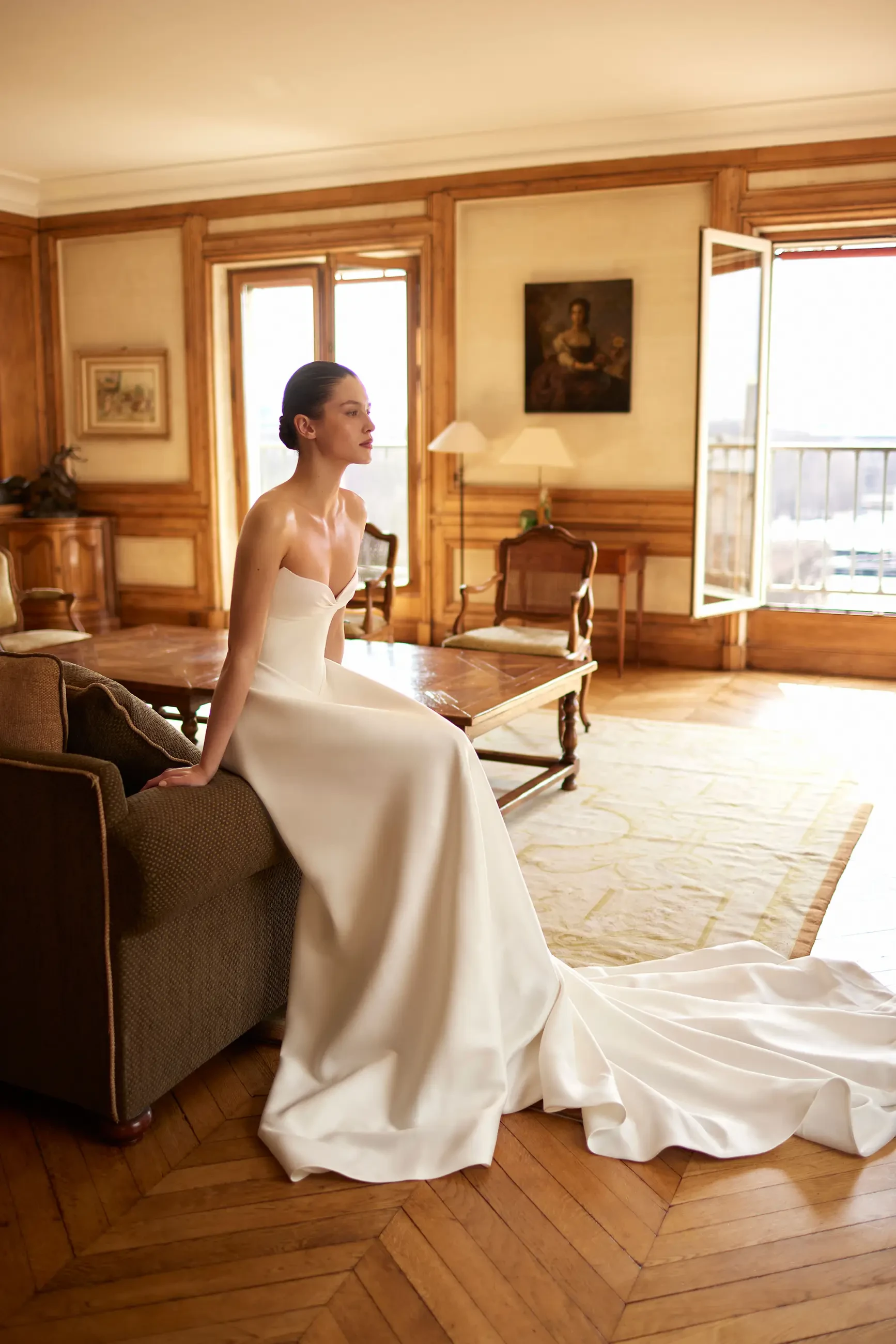 A woman in an elegant strapless white gown with a long train sits on a brown sofa in a classic, warmly-lit room with wooden decor and large windows.
