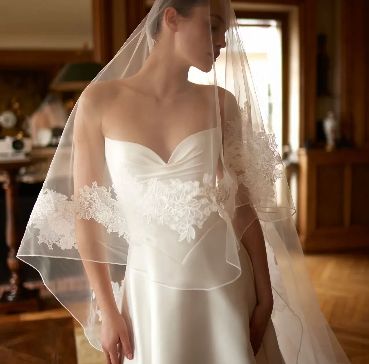 A bride stands gracefully in a satin wedding gown with a lace-embellished veil. She appears serene in a warmly lit, elegant room, conveying a sense of tranquility.