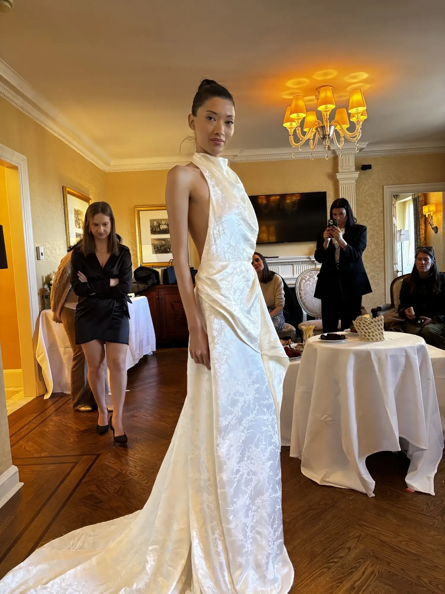 A model wearing a white, elegant dress stands confidently in a room filled with people and furniture.