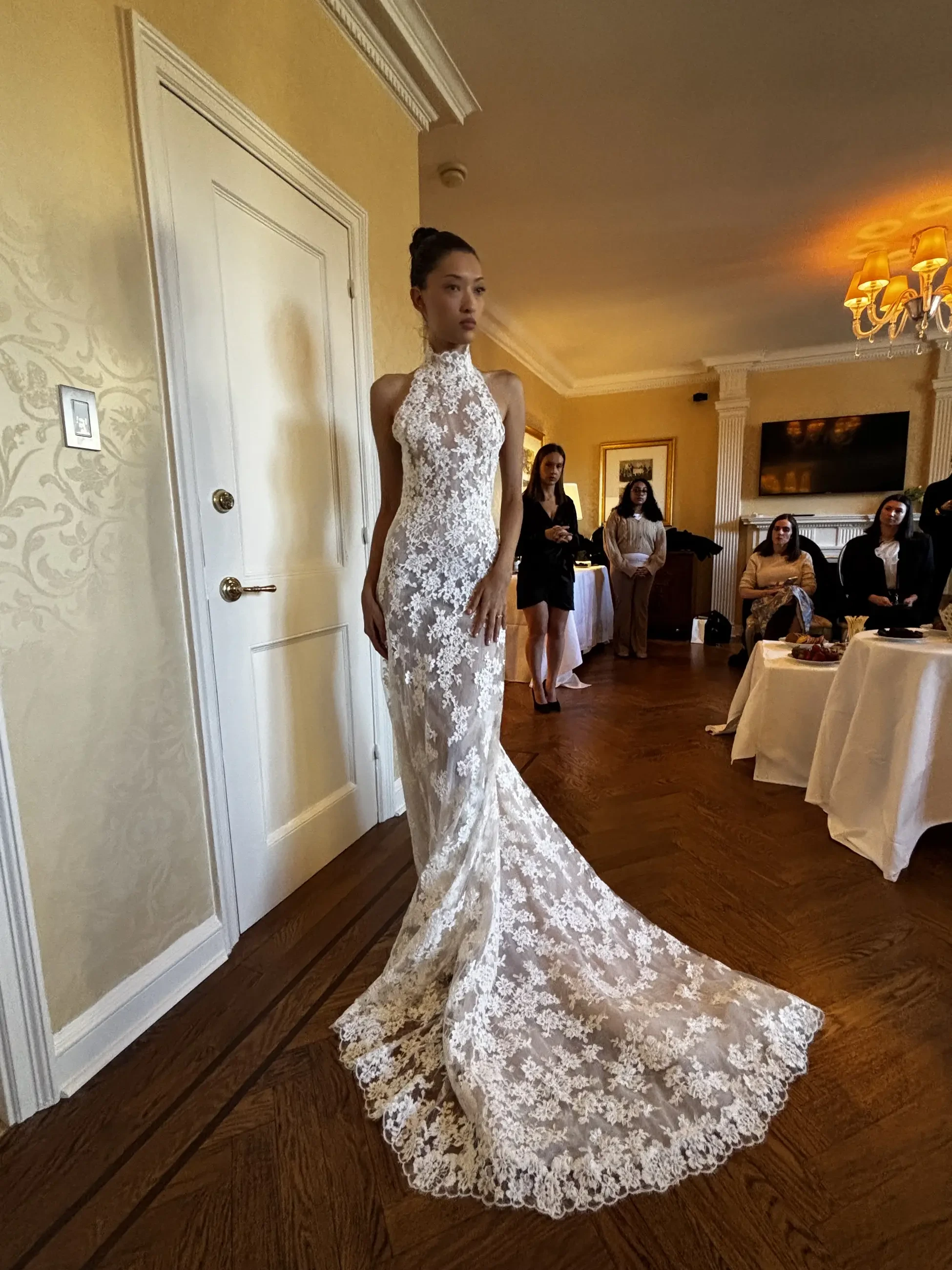 A model wearing an elegant white lace wedding dress poses in a room with soft lighting and several people in the background.
