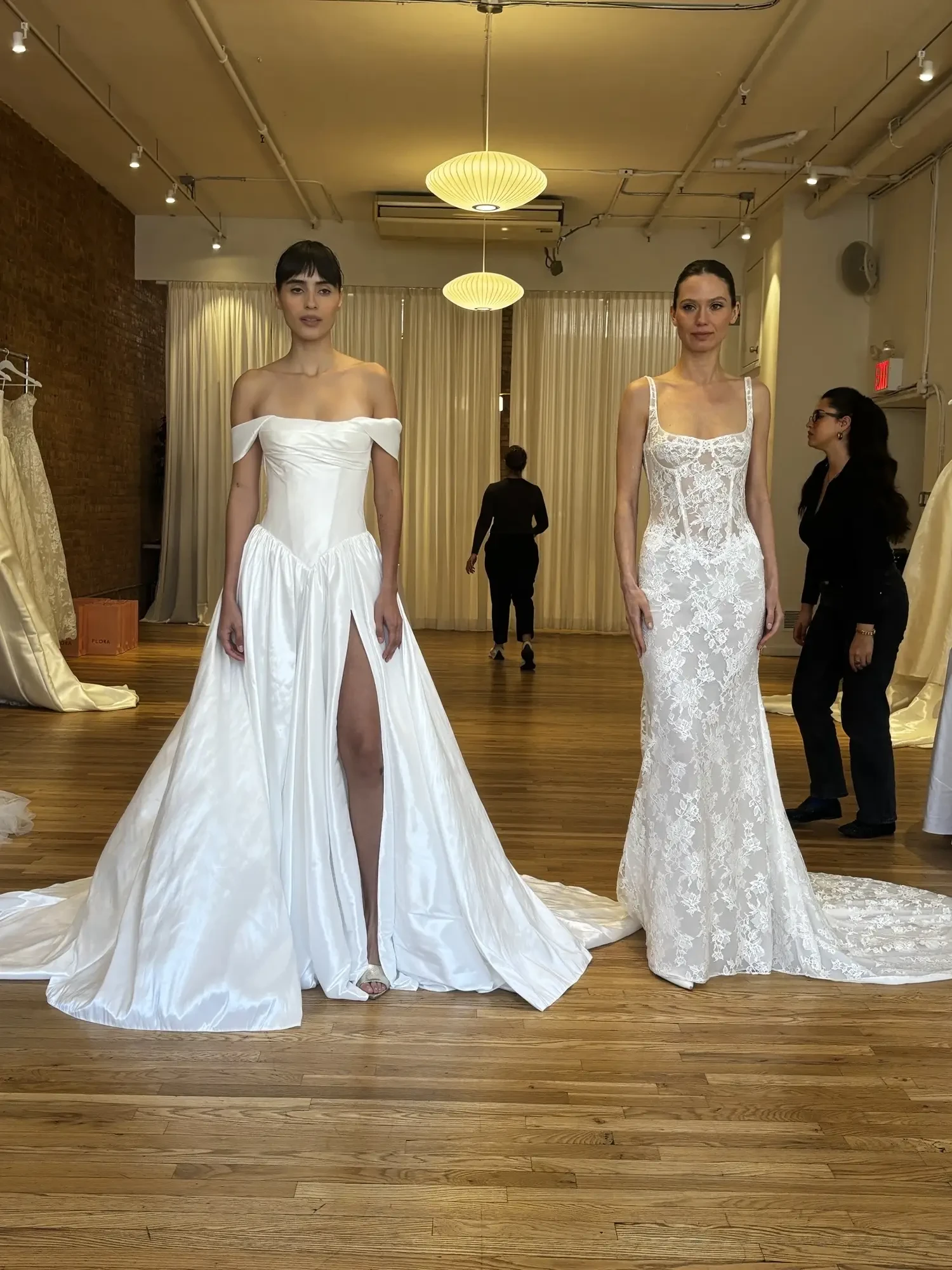 Two models wearing elegant wedding dresses in a spacious, well-lit room with wooden floors and fabric drapes. One dress is a white, off-the-shoulder gown with a high slit, and the other is a fitted lace gown with intricate details. A few people are present in the background.