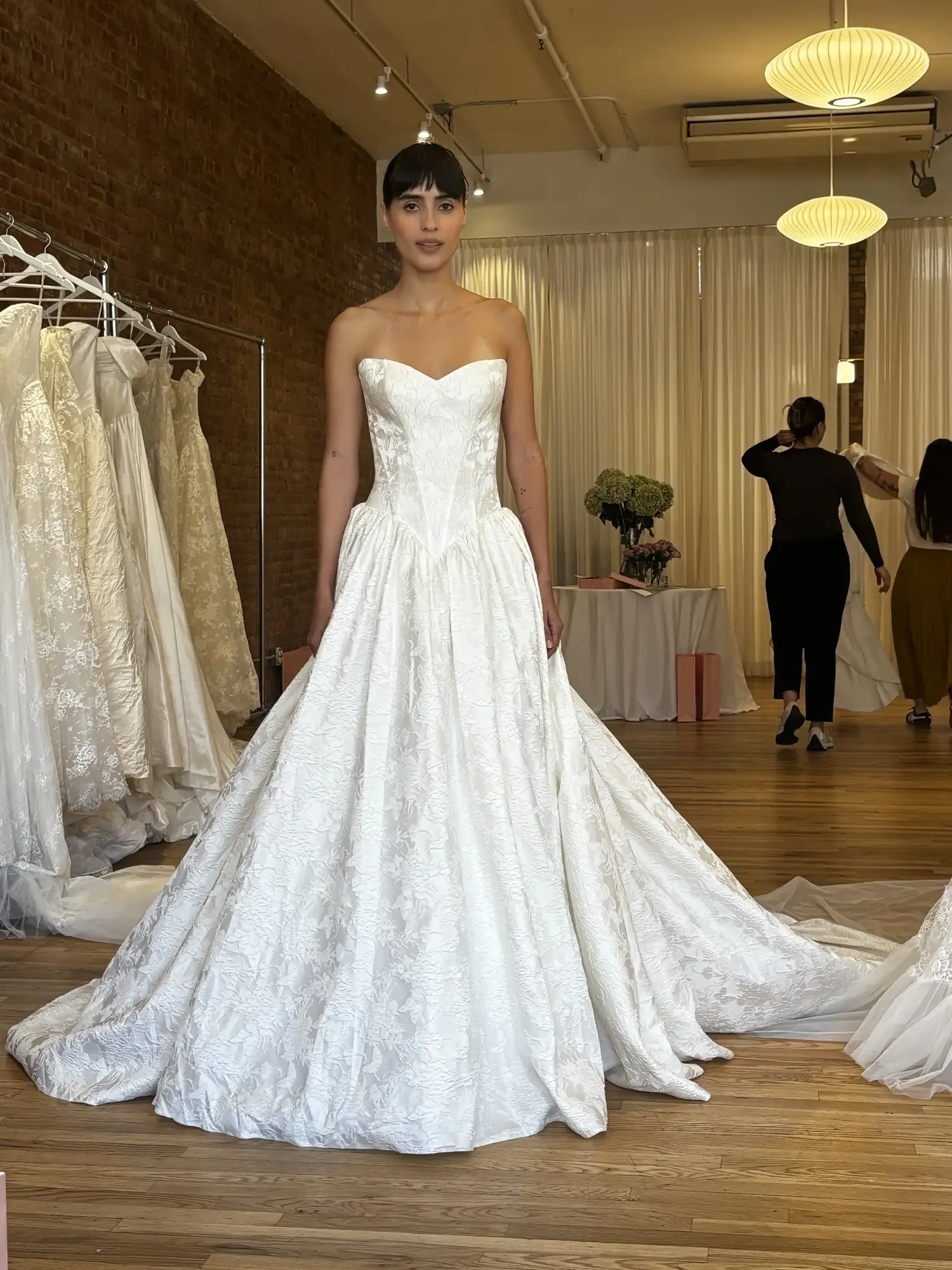 A woman in a strapless, white wedding dress stands in a bridal shop with other dresses visible in the background.