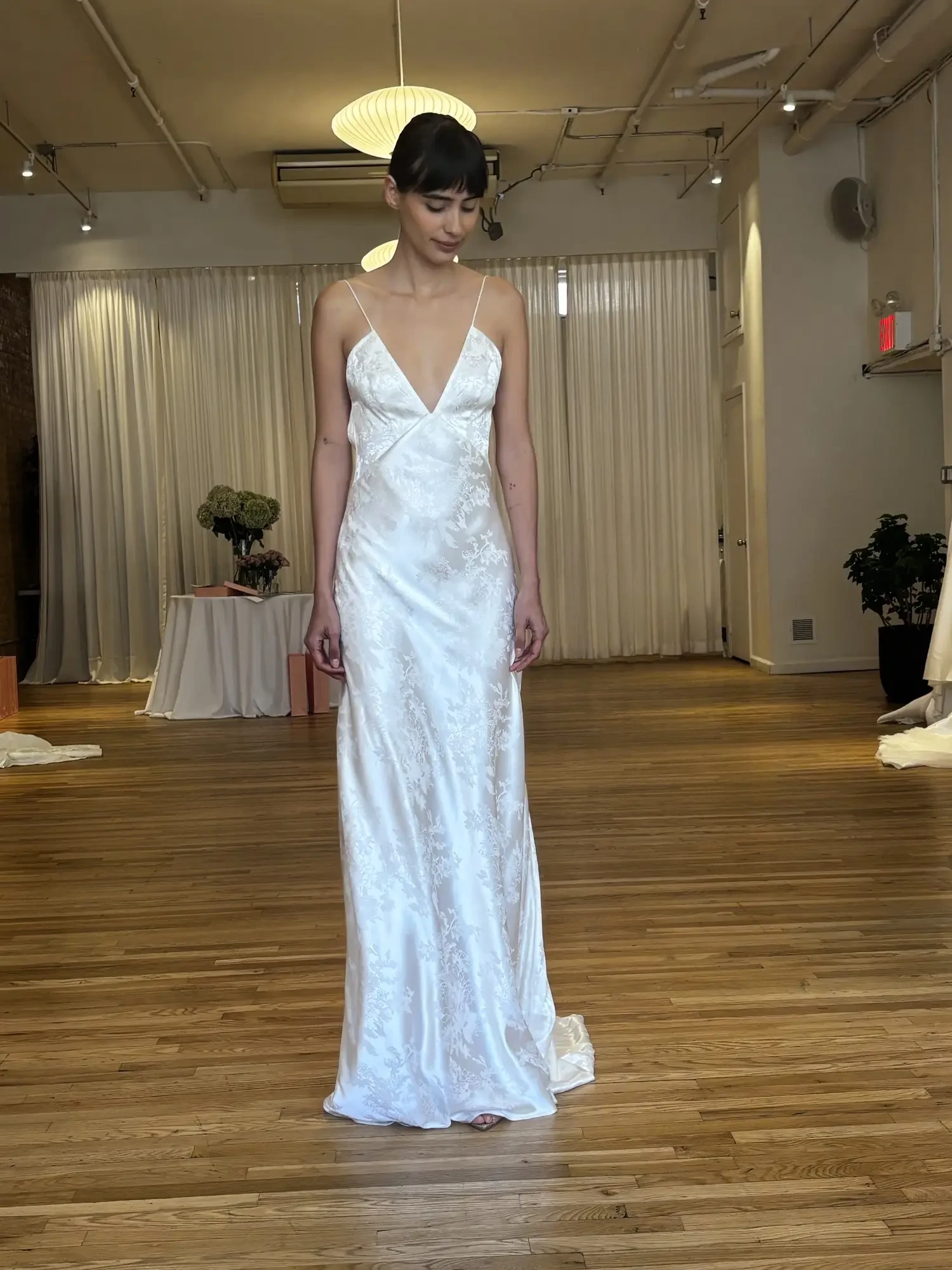 A model wearing a white satin gown with thin straps is standing in an empty room with wooden floors and soft lighting.
