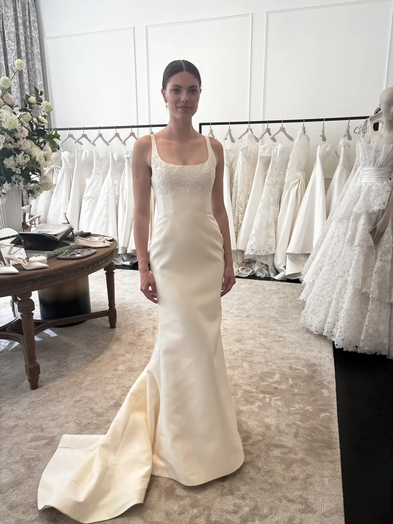 A model wearing an elegant white wedding dress stands in a bridal shop, with various gowns hanging in the background.