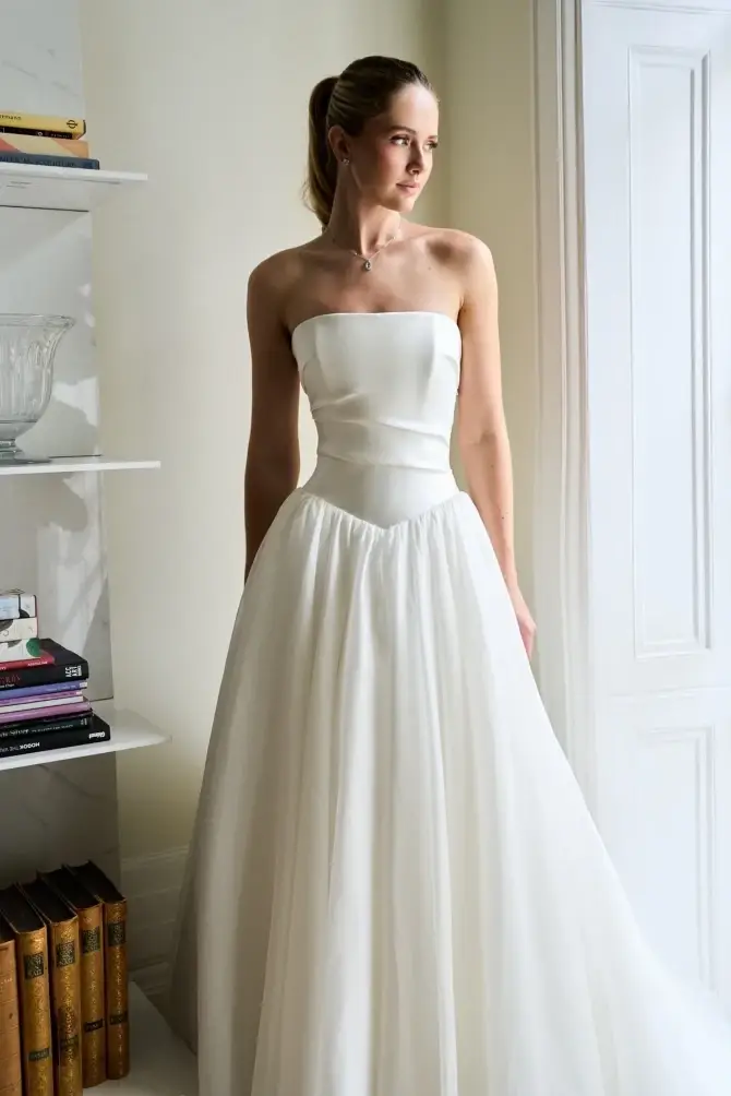 A woman in a strapless white gown stands elegantly in front of a bookshelf, looking off to the side.