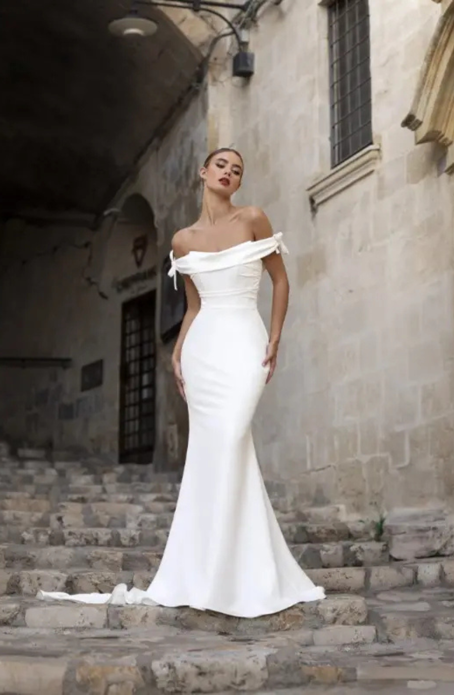 Model is wearing a minimalist off the shoulder fit and flare bridal gown with two delicate bows on each shoulder by valentini spose