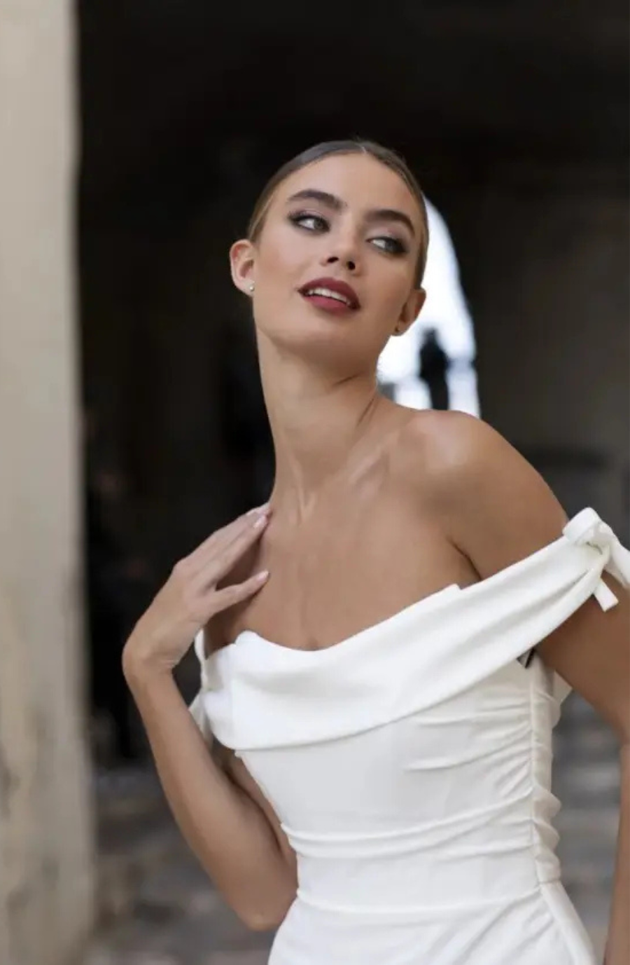 Model is wearing a minimalist off the shoulder fit and flare bridal gown with two delicate bows on each shoulder by valentini spose