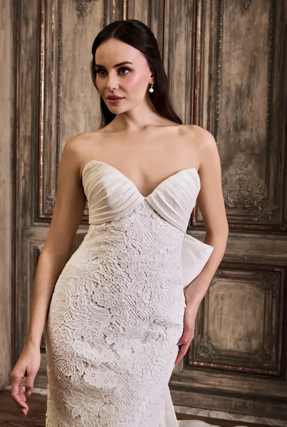 A woman in a strapless white dress with intricate lace details, standing against a vintage backdrop.