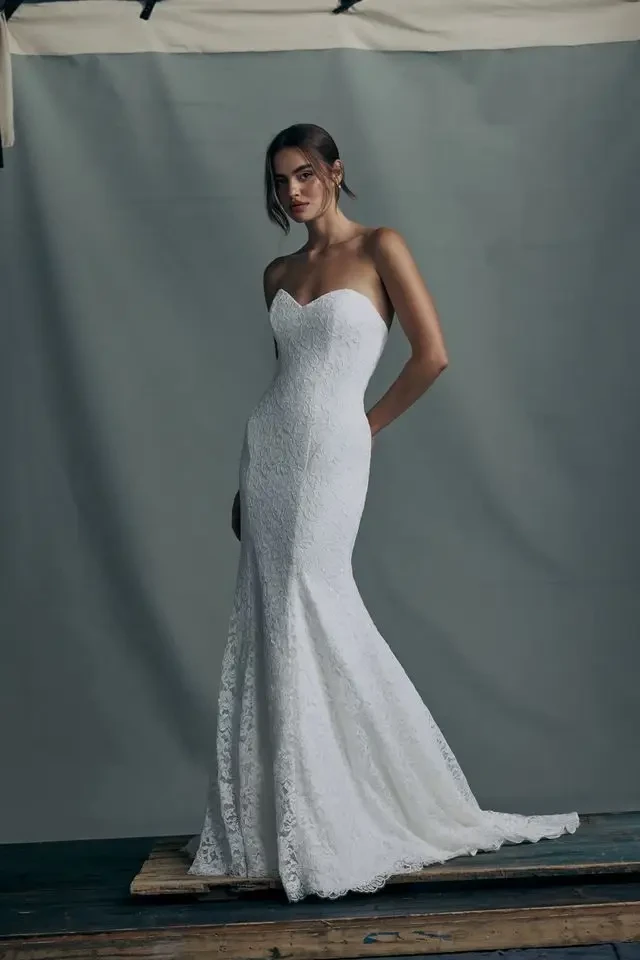 A model wearing a strapless white lace wedding gown, standing in front of a neutral backdrop.