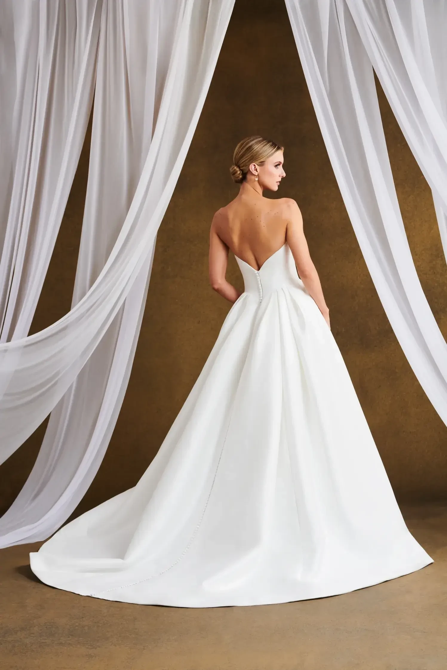 Model is wearing a Mikado ball gown with a scooped neckline, curved basque waist, pleated skirt, and covered buttons.