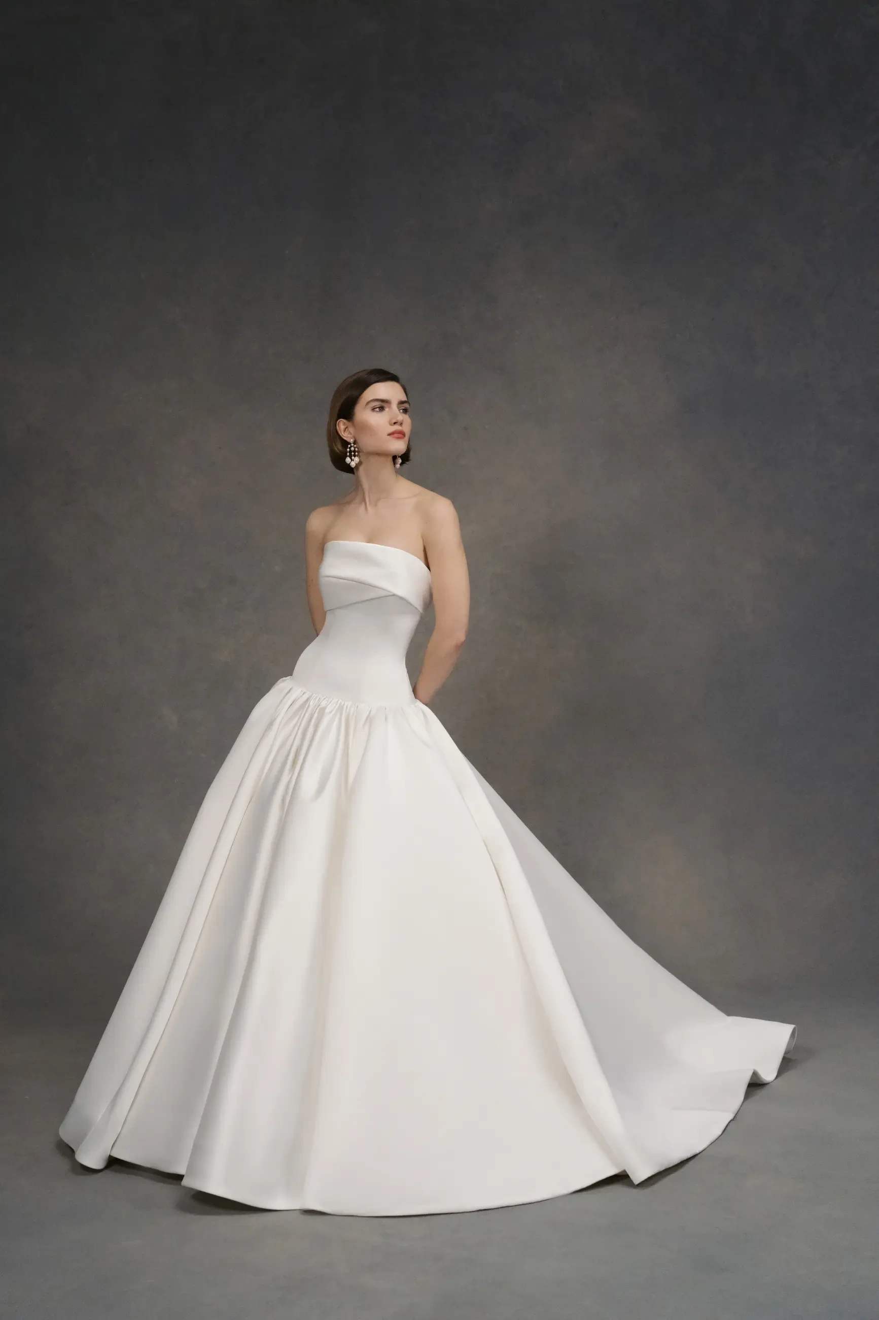 A woman in an elegant, strapless white ball gown stands against a textured grey backdrop. Her expression is poised and serene, exuding grace.