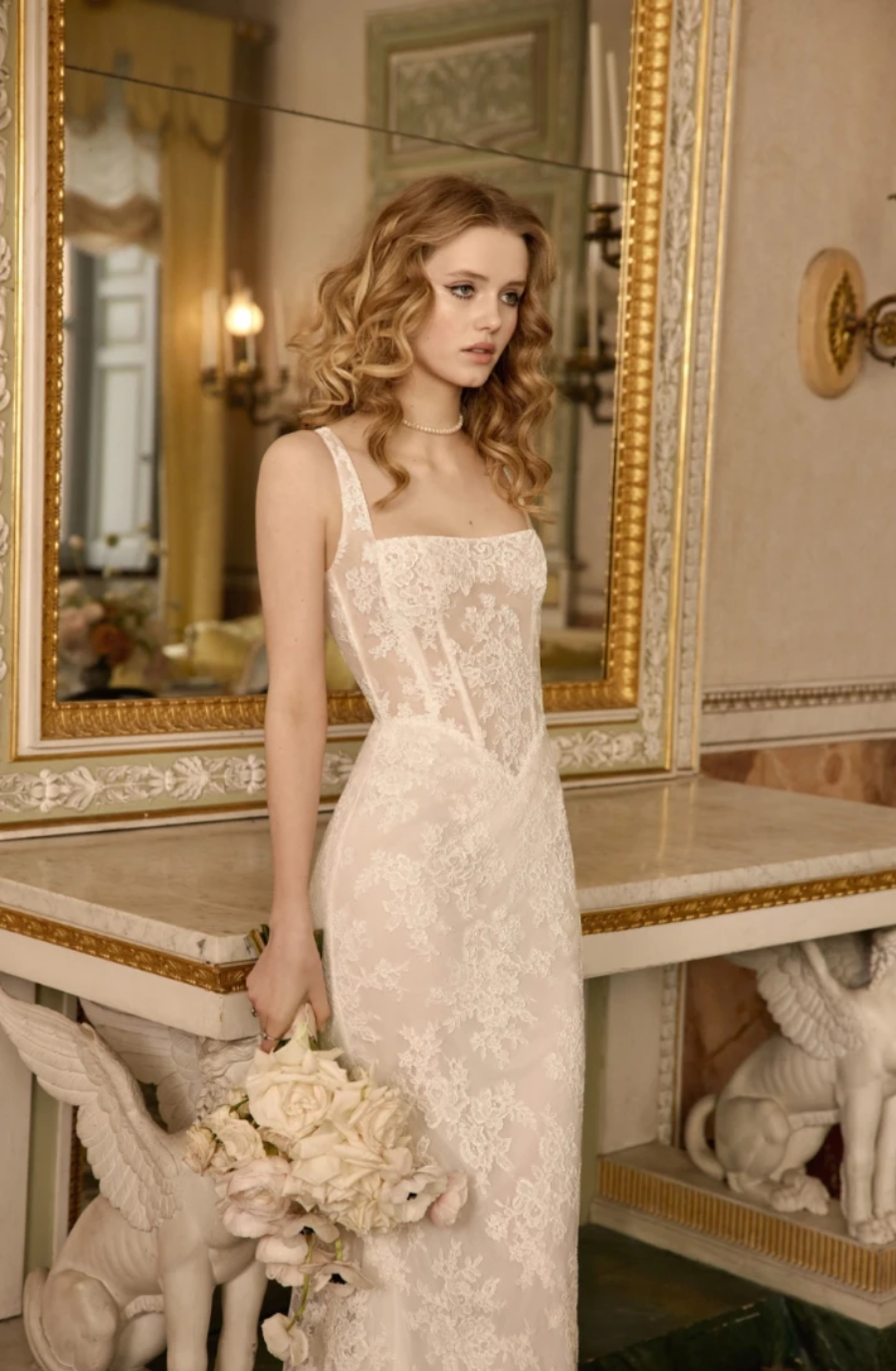 A woman in an elegant, lace wedding dress stands by a marble table, holding a bouquet of flowers, with ornate decor and mirrors in the background.