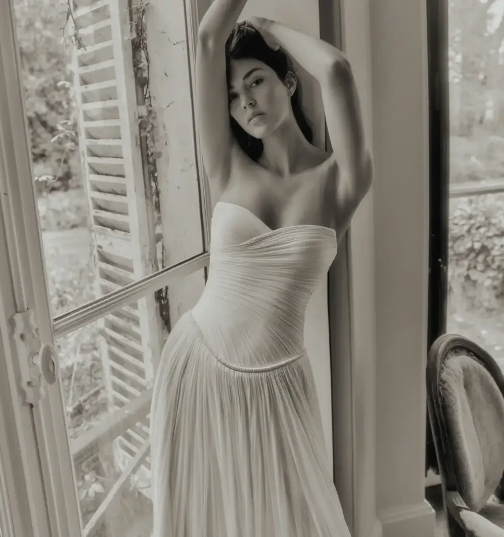 A woman in a strapless, flowing dress leaning against a window, with her arms raised above her head, in a monochrome setting.