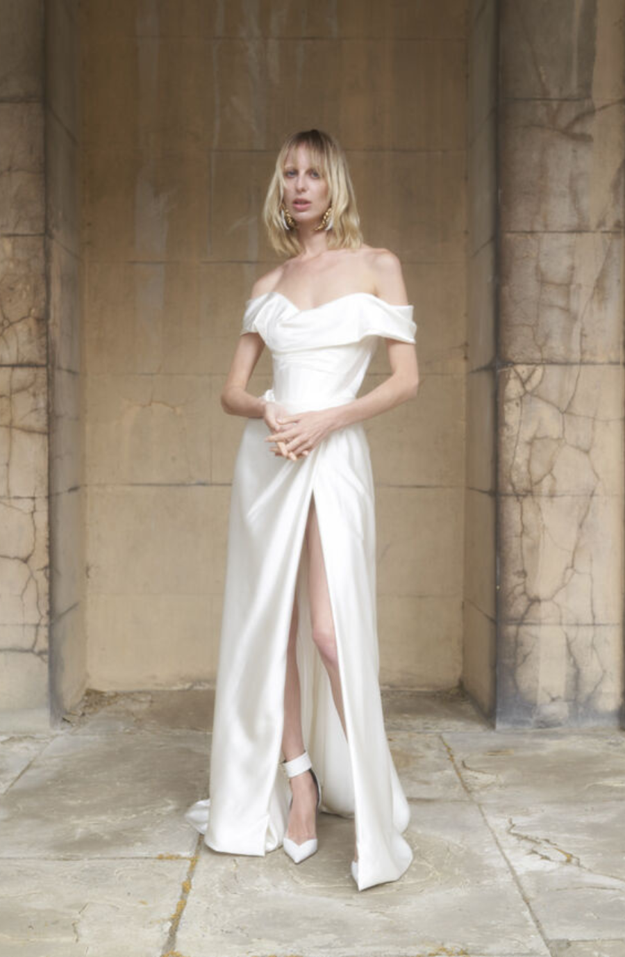A woman stands confidently in an elegant, off-shoulder white gown with a thigh-high slit, set against a rustic stone backdrop, conveying sophistication.