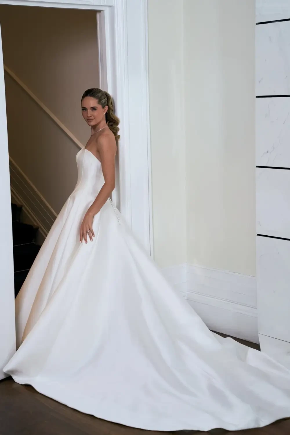 A bride in a white wedding dress stands near a doorway, featuring a strapless design and a long train.