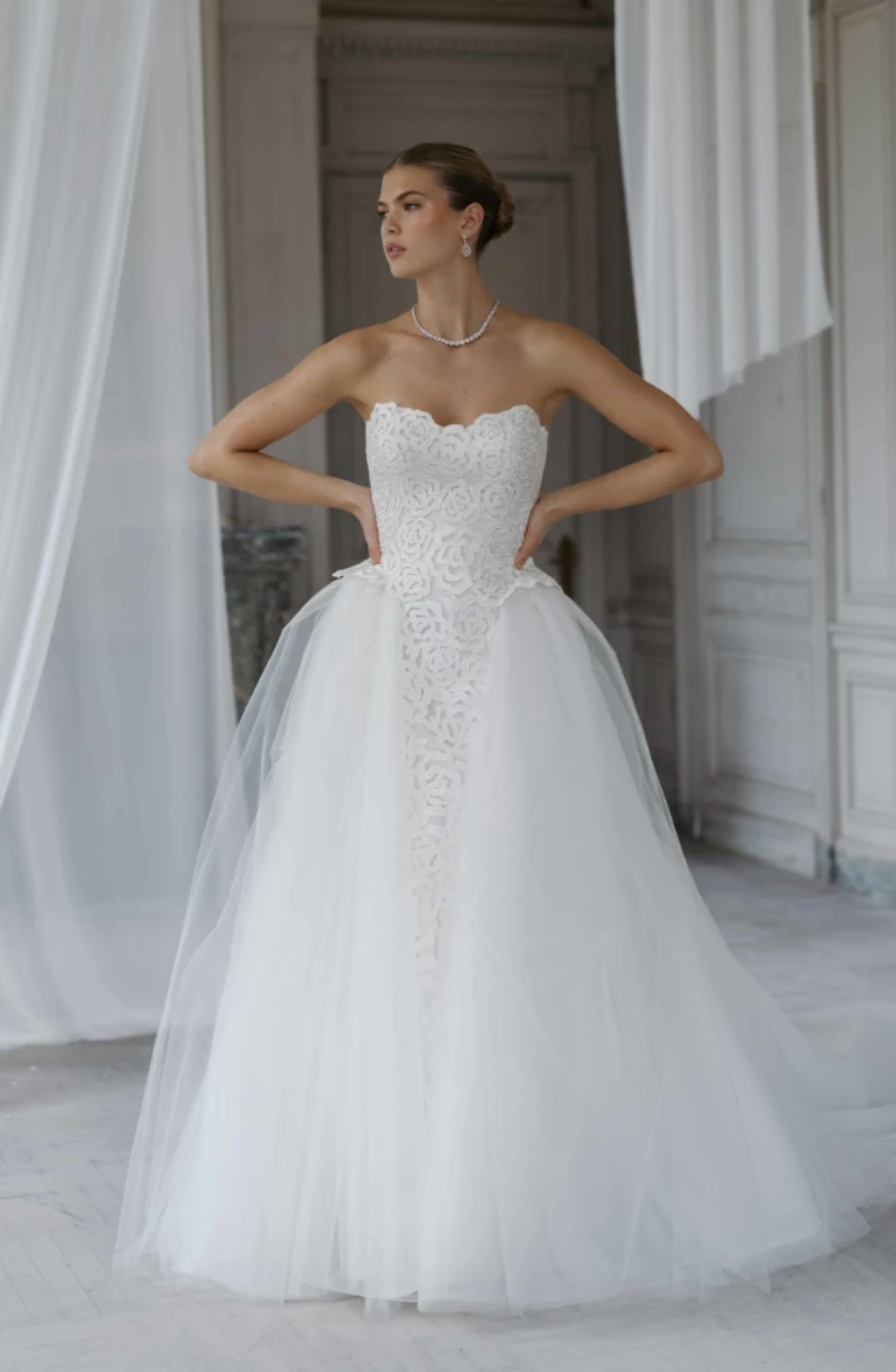 A woman in a strapless white bridal gown with a tulle skirt and intricate lace detailing, standing in an elegant indoor setting.