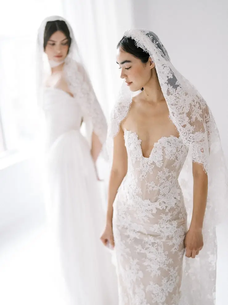 Two women in elegant wedding dresses, one wearing a sheer veil and the other with a flowing gown, posing indoors with natural light.
