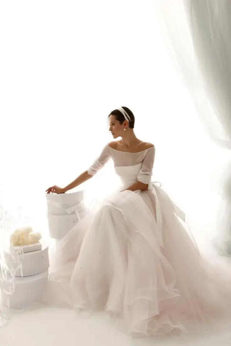 A woman in an elegant, off-shoulder wedding dress sitting near white gift boxes, with a soft, light background.
