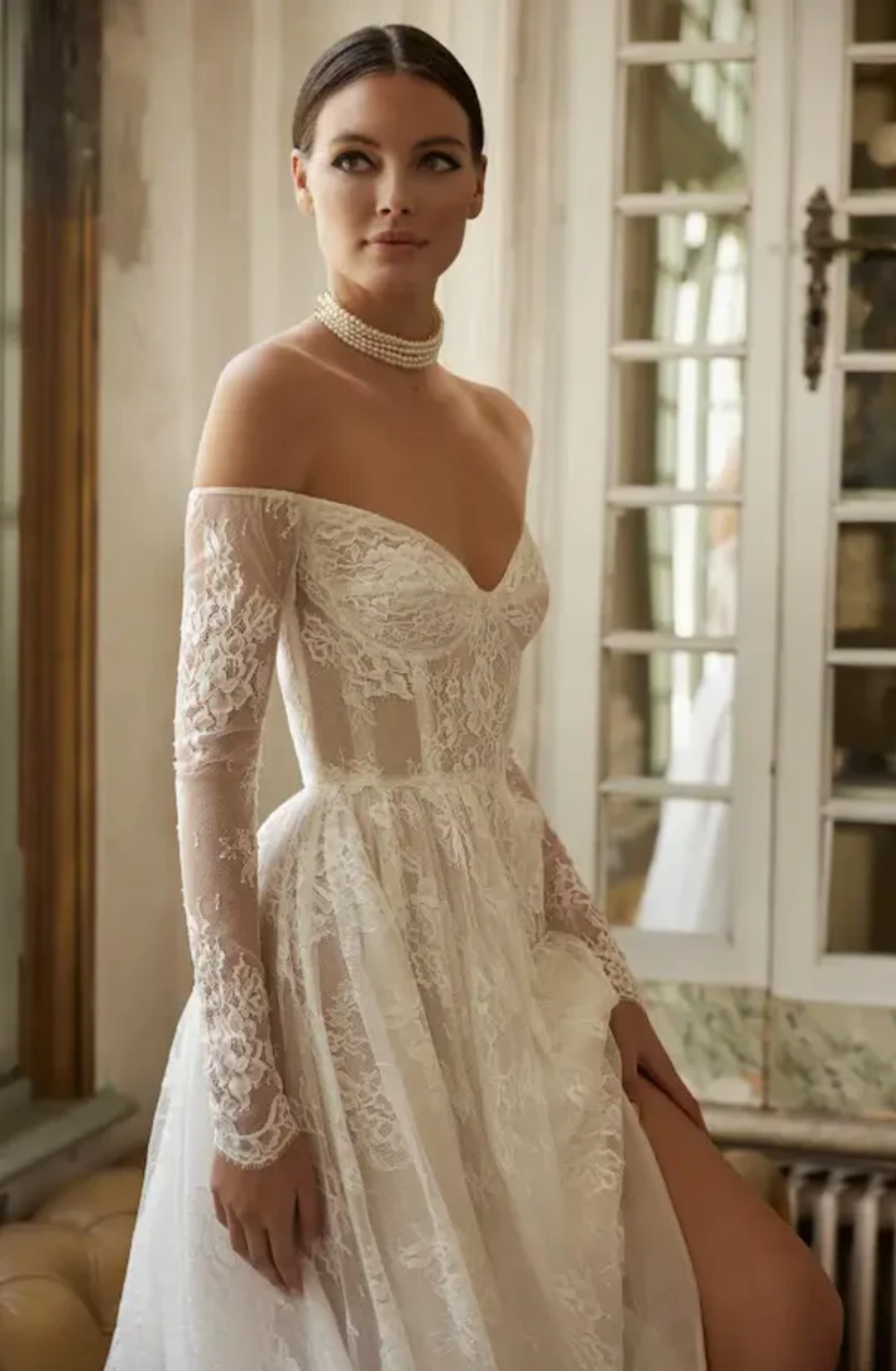 A woman wearing an elegant off-the-shoulder white lace wedding dress with long sleeves, standing in a softly lit room.