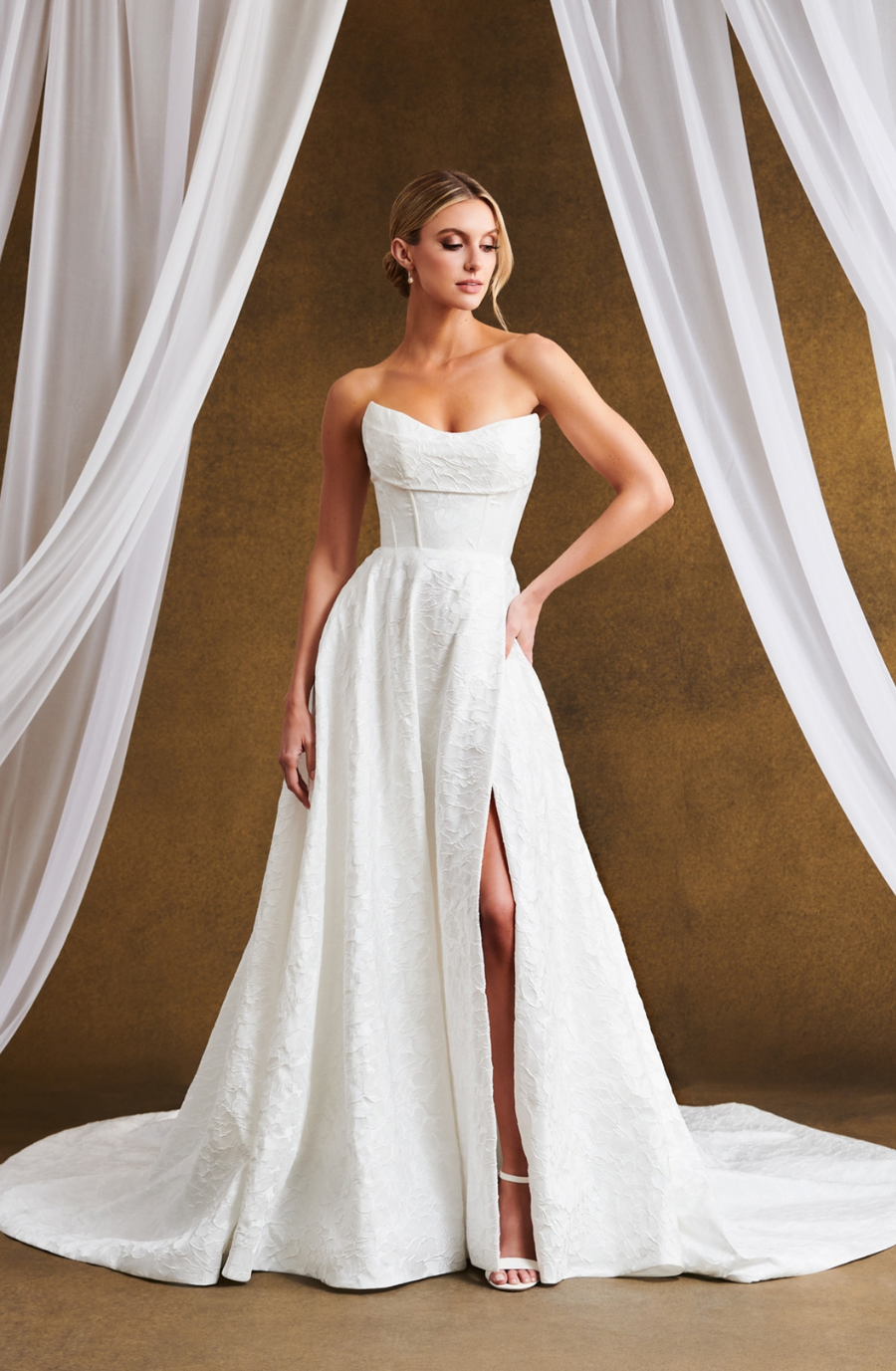 Bridal model in a strapless white gown with a thigh-high slit, standing on a gold backdrop. Draped white fabric adds elegance and sophistication.