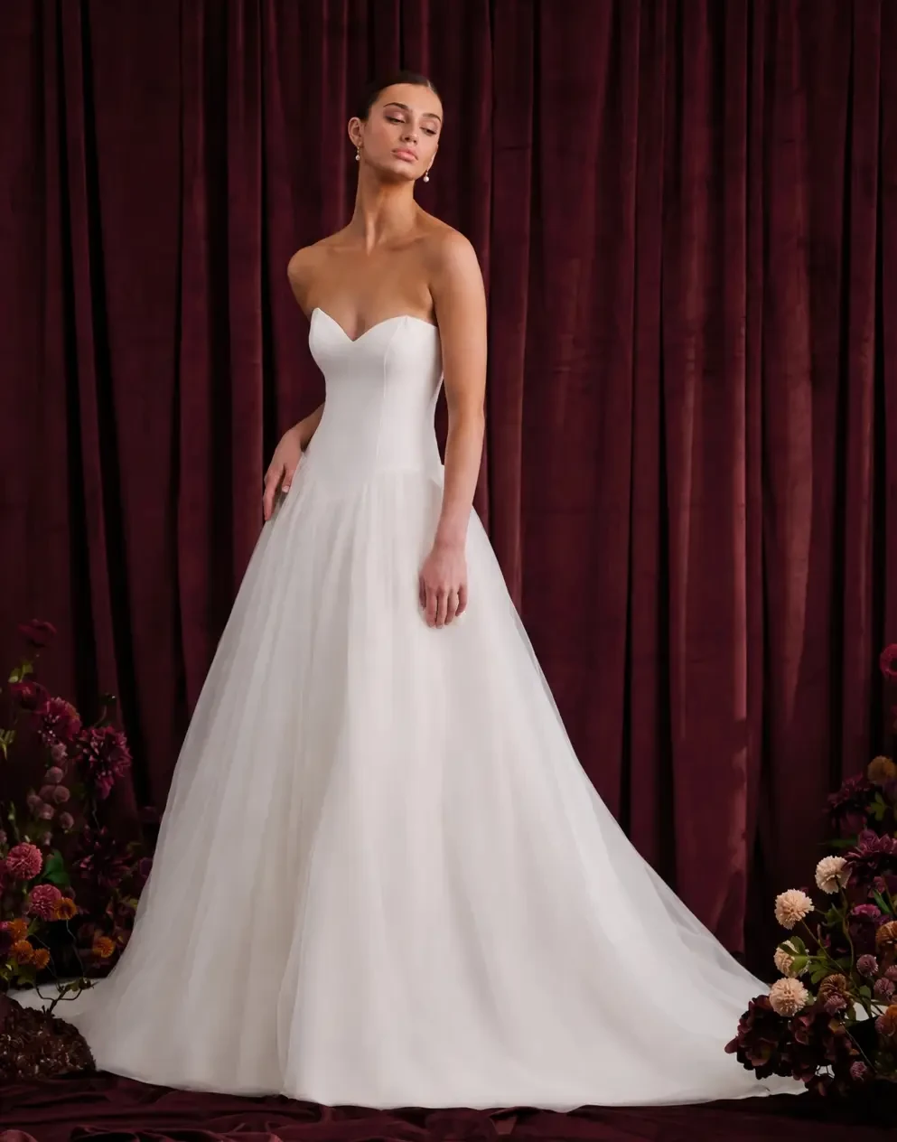 A model wearing a strapless white wedding gown, standing against a deep red backdrop with floral arrangements at her feet.