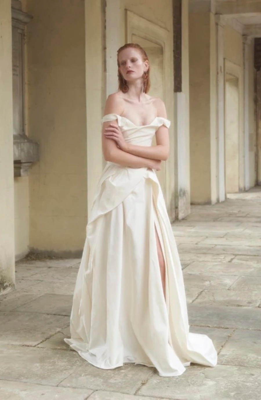 A woman stands confidently in an off-the-shoulder, cream-colored gown with a thigh-high slit. The setting is an elegant, historical hallway, evoking a timeless and serene atmosphere.