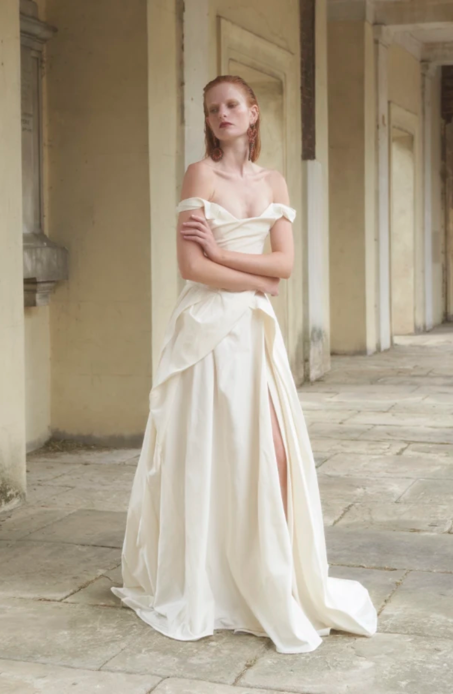 A model wearing an off-shoulder white dress stands in a corridor with classical architecture. The dress features a high slit and flowing fabric, complemented by the model's natural makeup and hairstyle.