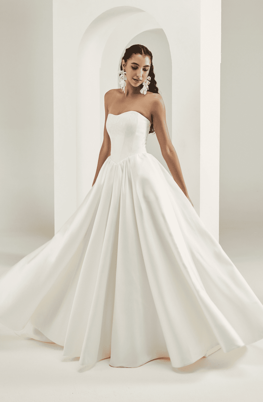 A woman in an elegant strapless wedding gown stands gracefully, with a full, flowing skirt. Her hair is styled back and she wears large, ornate earrings.